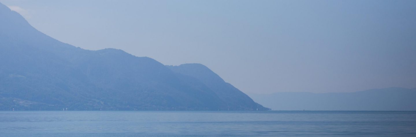 Tranquil blue lake landscape with soft water ripples and misty mountains under a pale sky.