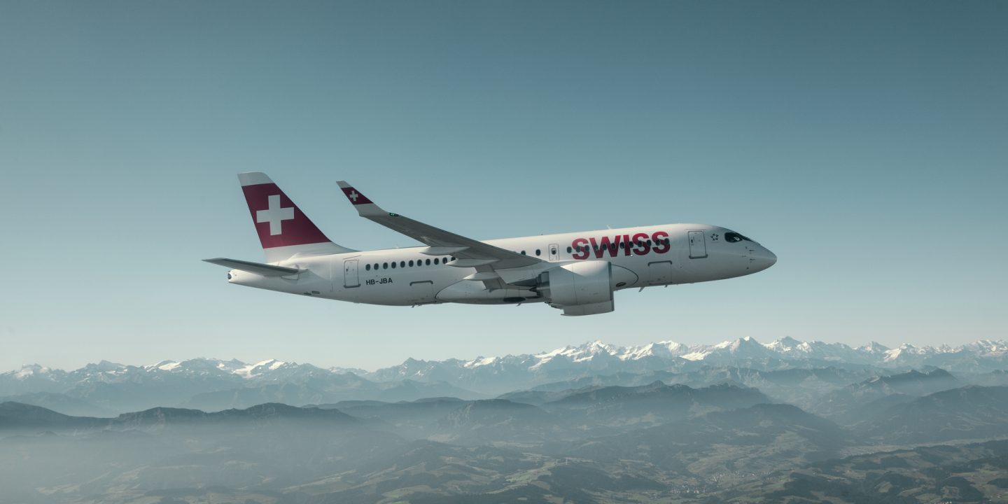 SWISS aircraft flying high above snow-capped mountains & green valleys under blue sky.