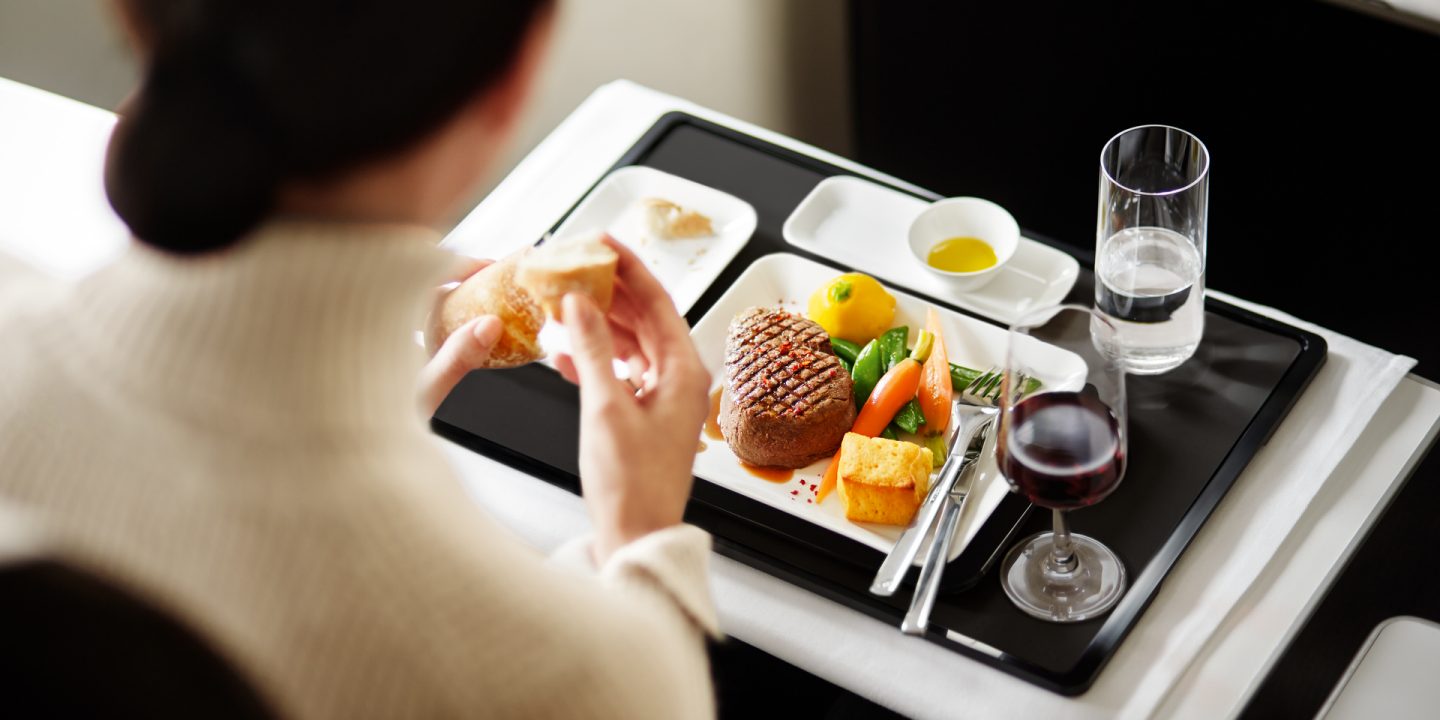 Passenger enjoying premium in-flight meal: steak, vegetables, bread, wine.