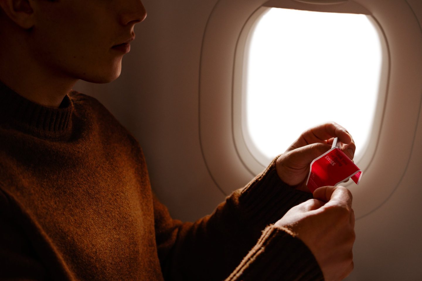 Passagier im braunen Pullover, der an einem Flugzeugfenster eine rote Schweizer Schokoladenverpackung öffnet.