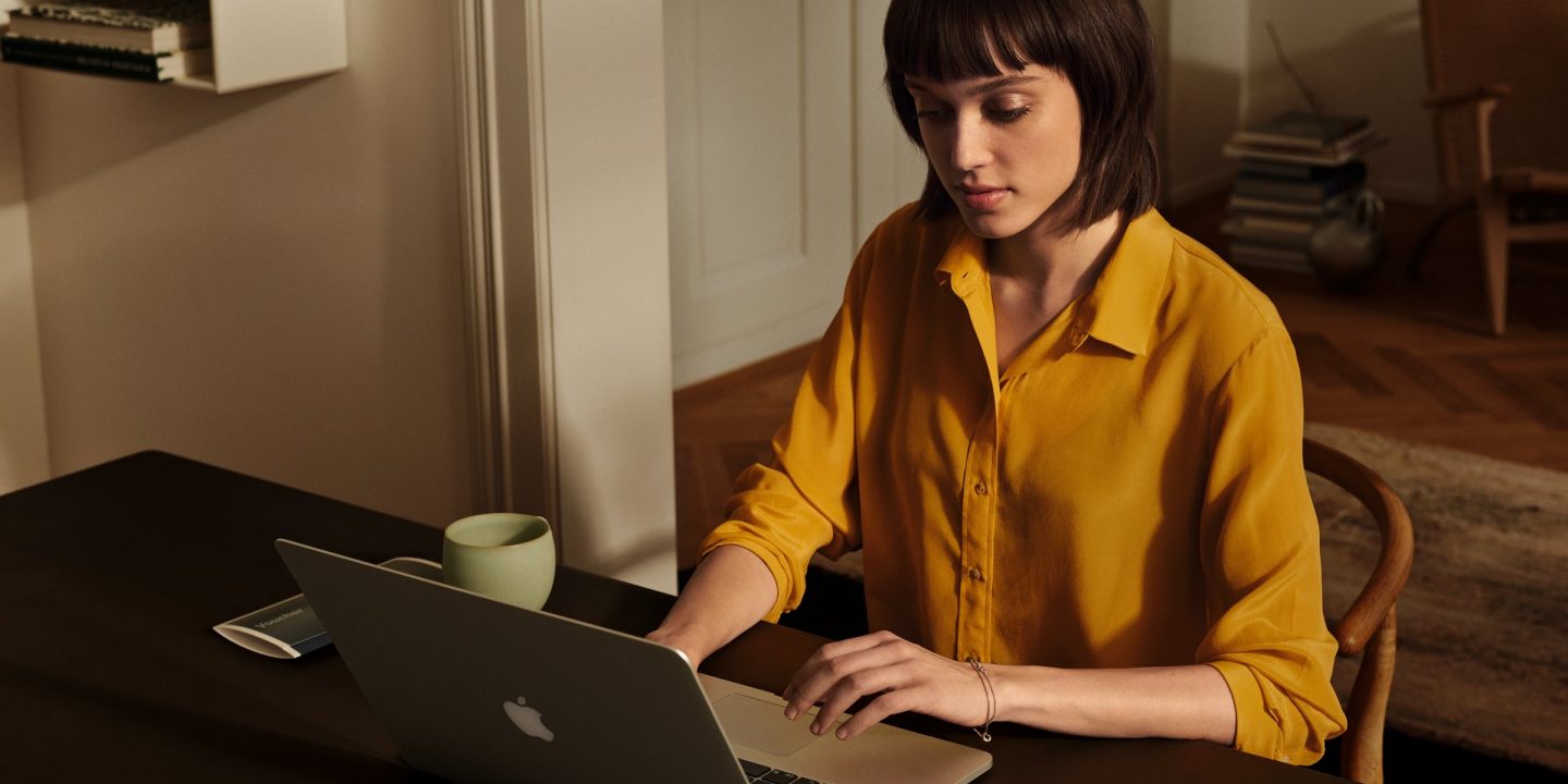 A woman in a yellow shirt sitting at a desk and working on a laptop.