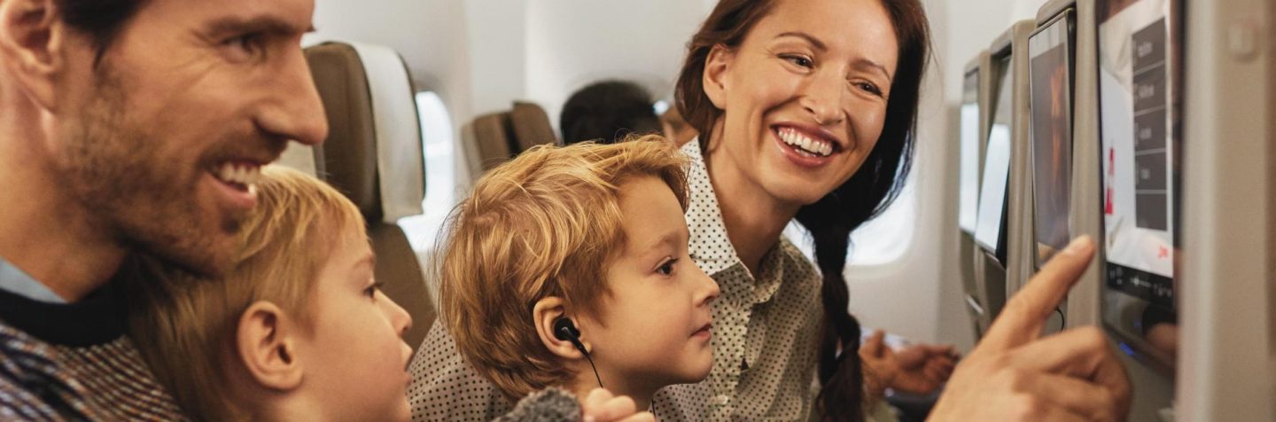 A family seated on an airplane, with a screen displaying the flight time in the background.