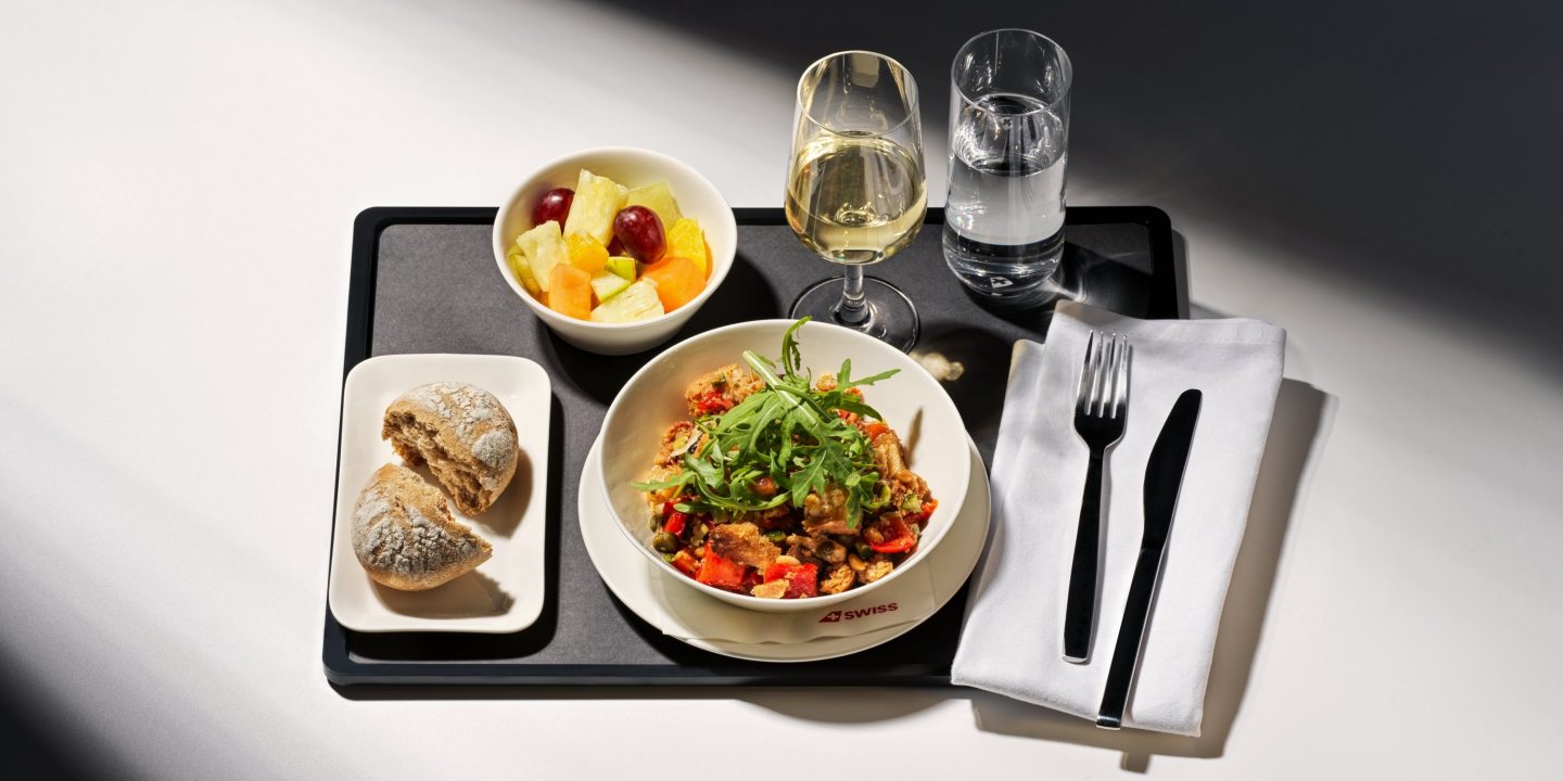 Vibrant SWISS in-flight meal: main dish with arugula, fresh fruit, bread, wine, and water.