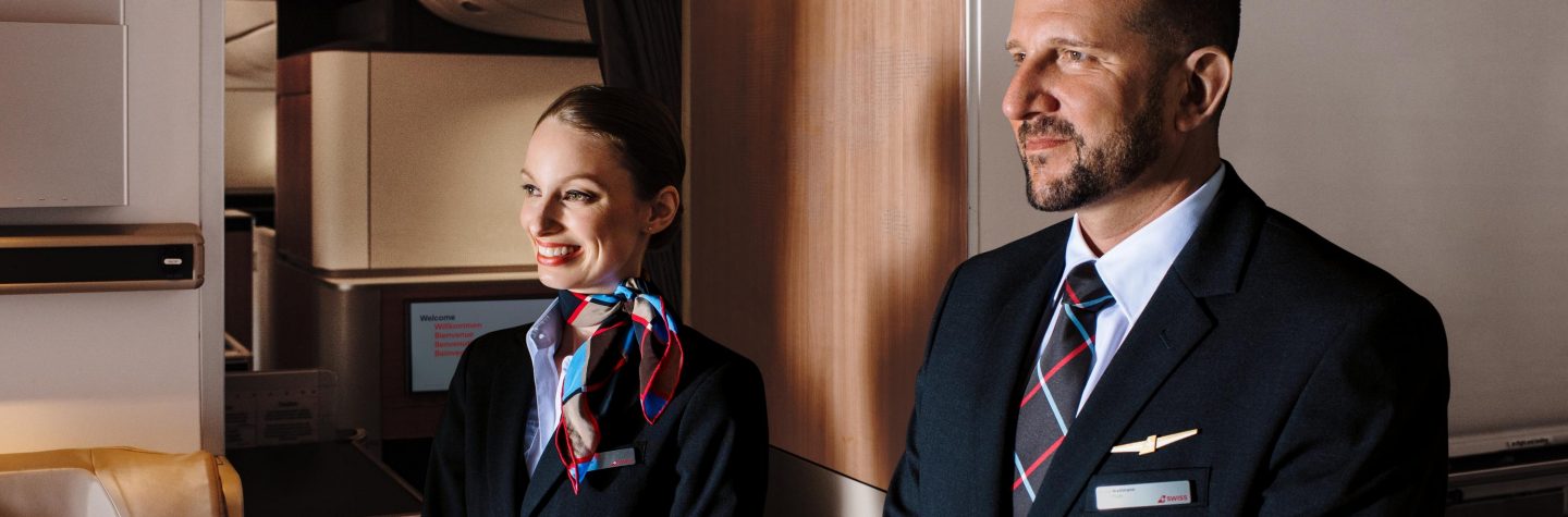 A man and woman in business attire stand together in front of a mirror, reflecting a professional setting.