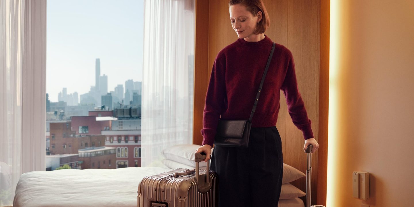 A woman with luggage stands by a window, preparing for an overnight hotel stay.