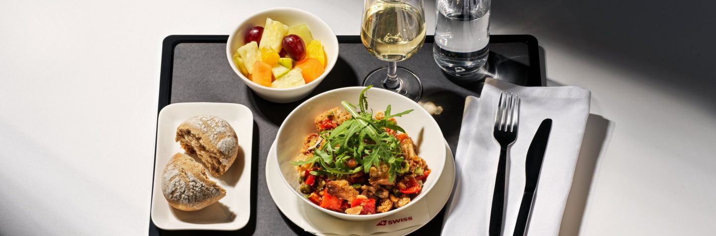 A gourmet inflight meal featuring a fresh salad, bread rolls & fruit salad served on a tray with a glass of white wine.