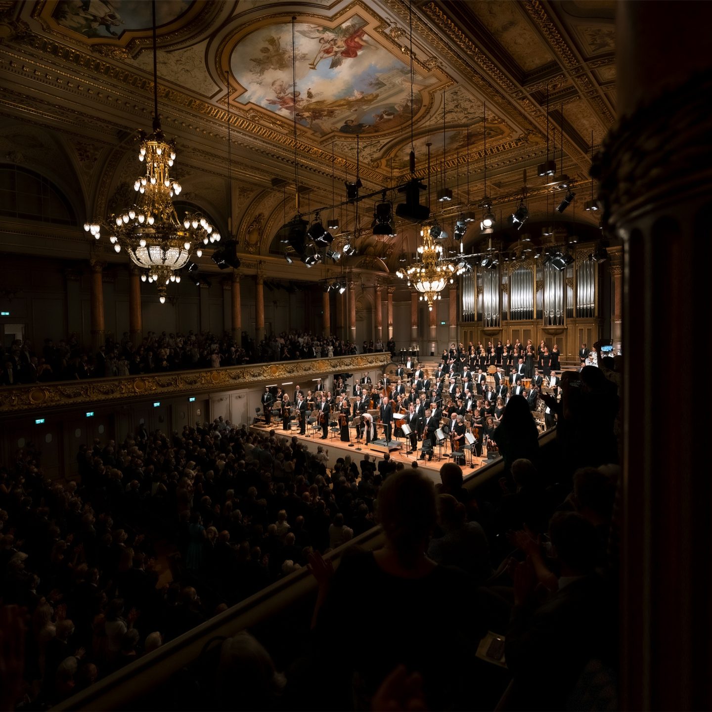 Orchestra and choir bowing on stage in grand concert hall with ornate ceiling and chandeliers, as audience applauds.