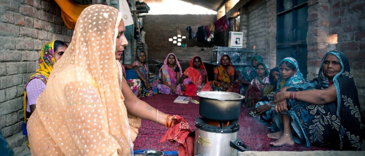 A woman in a sari is preparing a dish while a group of women watches with interest.