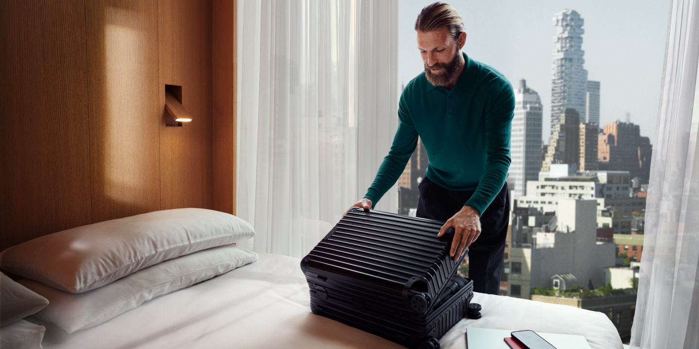 A man stands by a bed with a suitcase, preparing for travel in a cozy room.