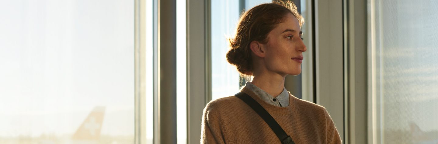 Person with a bun hairstyle and a brown sweater standing near large glass windows.