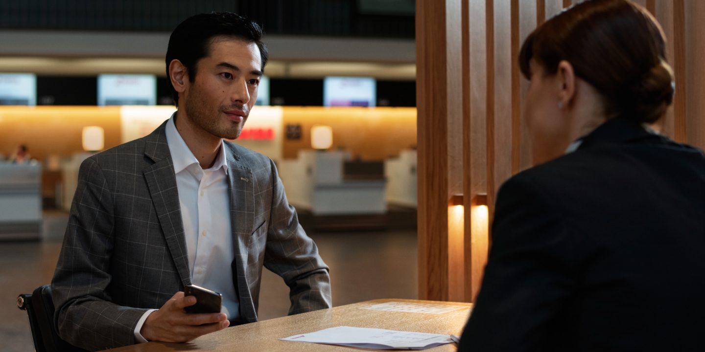 A man dressed in a suit sits at a desk, conversing on his cell phone with a serious expression.