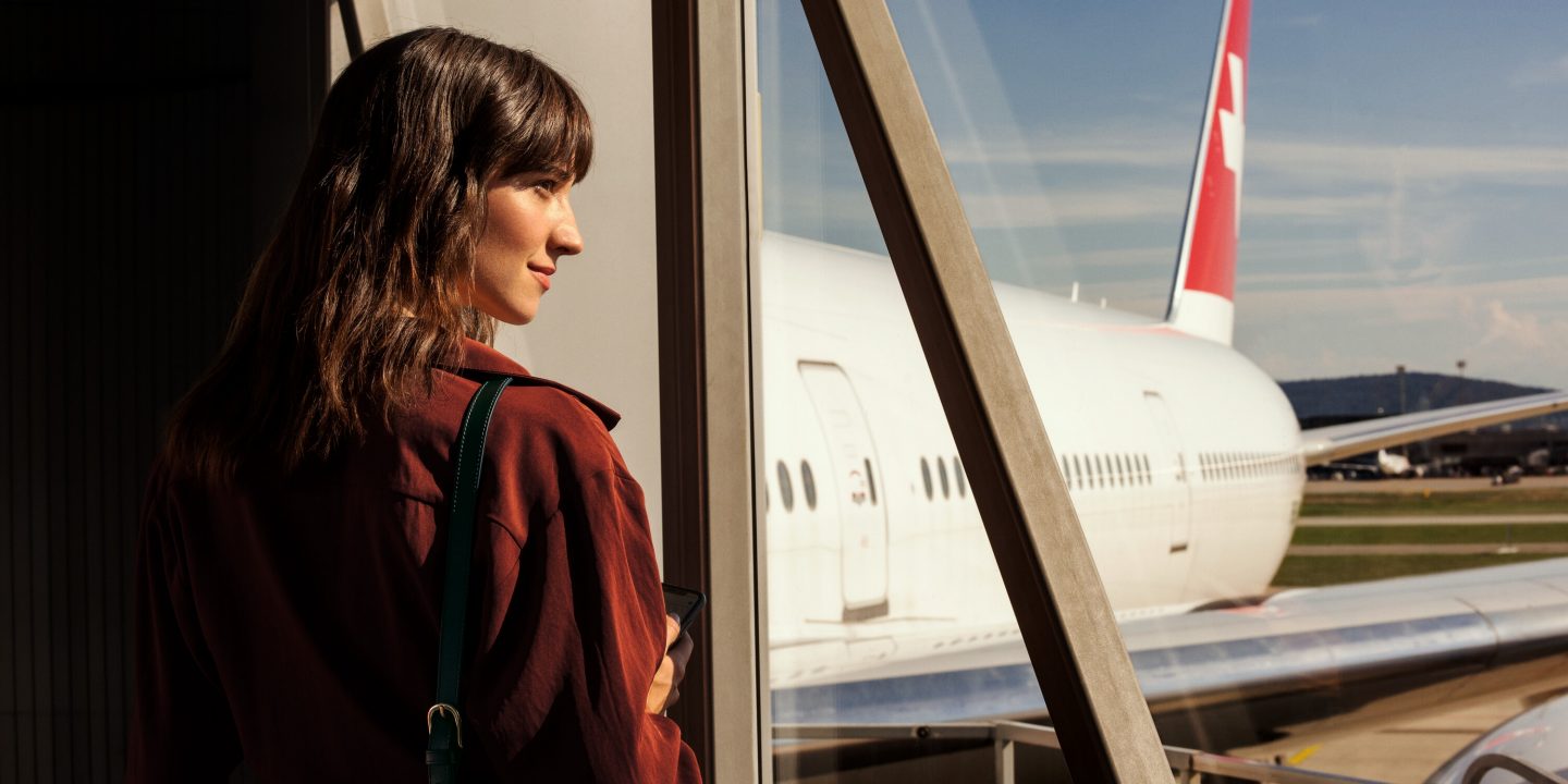 Smiling traveler at airport window, viewing a SWISS plane under sunny skies.