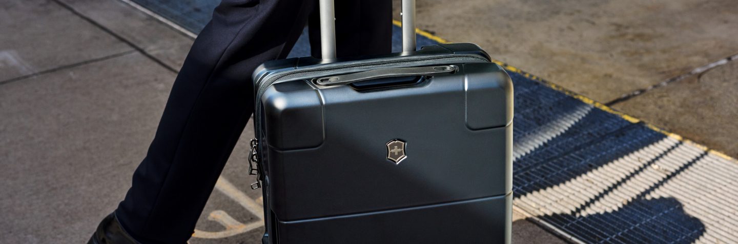 Person rolling a dark metallic suitcase on a paved surface.