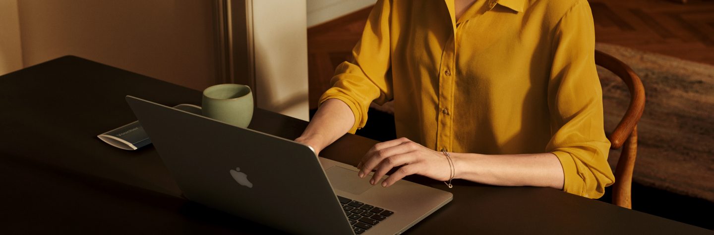 A woman in a yellow blouse sitting with her laptop at her desk; her head is cut away with the focus on her laptop